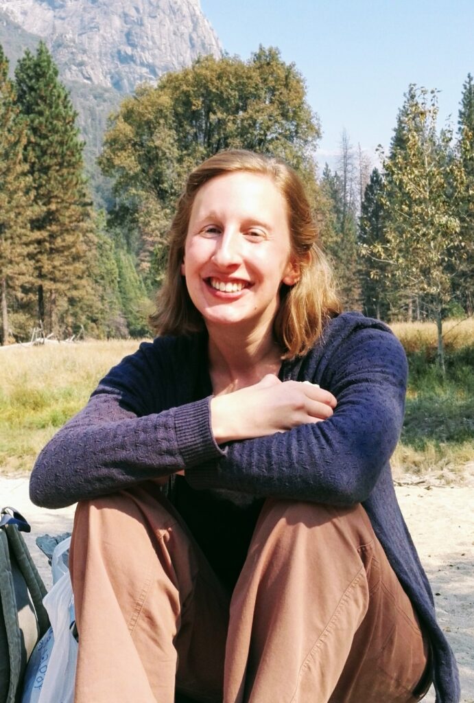 A photograph of a woman with chin length blond hair smiling at the camera. She is outside and has her arms crossed and resting on her knees.